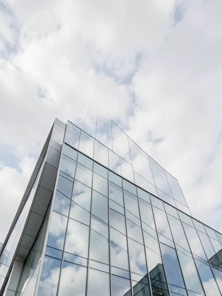 Modern glass office building exterior reflecting clouds, looking up from ground 