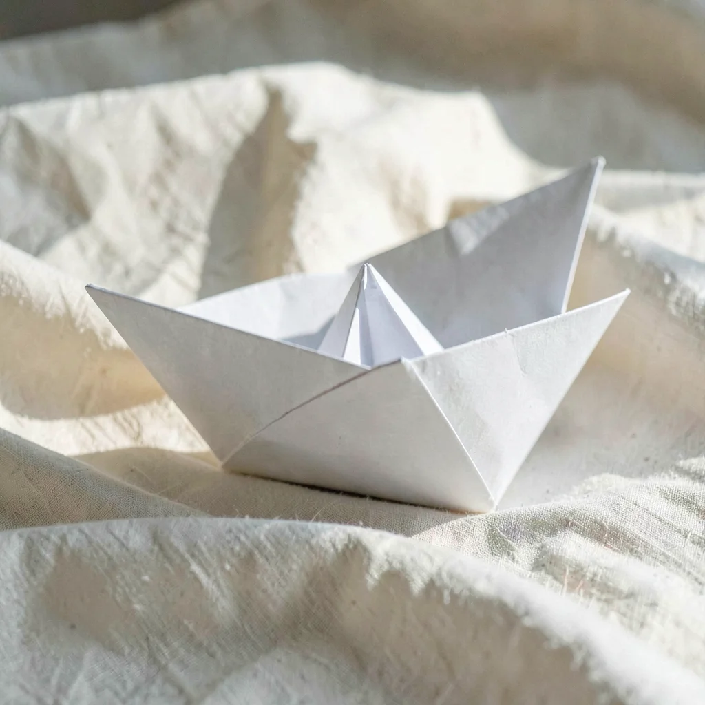 Folded paper boat resting on a bed of cream linen, sidelit, hairline crease catc