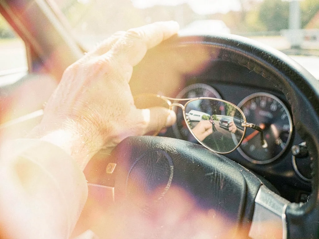 Man's weathered hand resting on a car steering wheel, dashboard reflected in avi