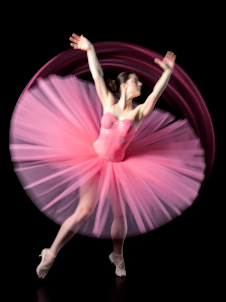 Ballet dancer mid-pirouette creating spiral blur of pink tulle and pale skin aga