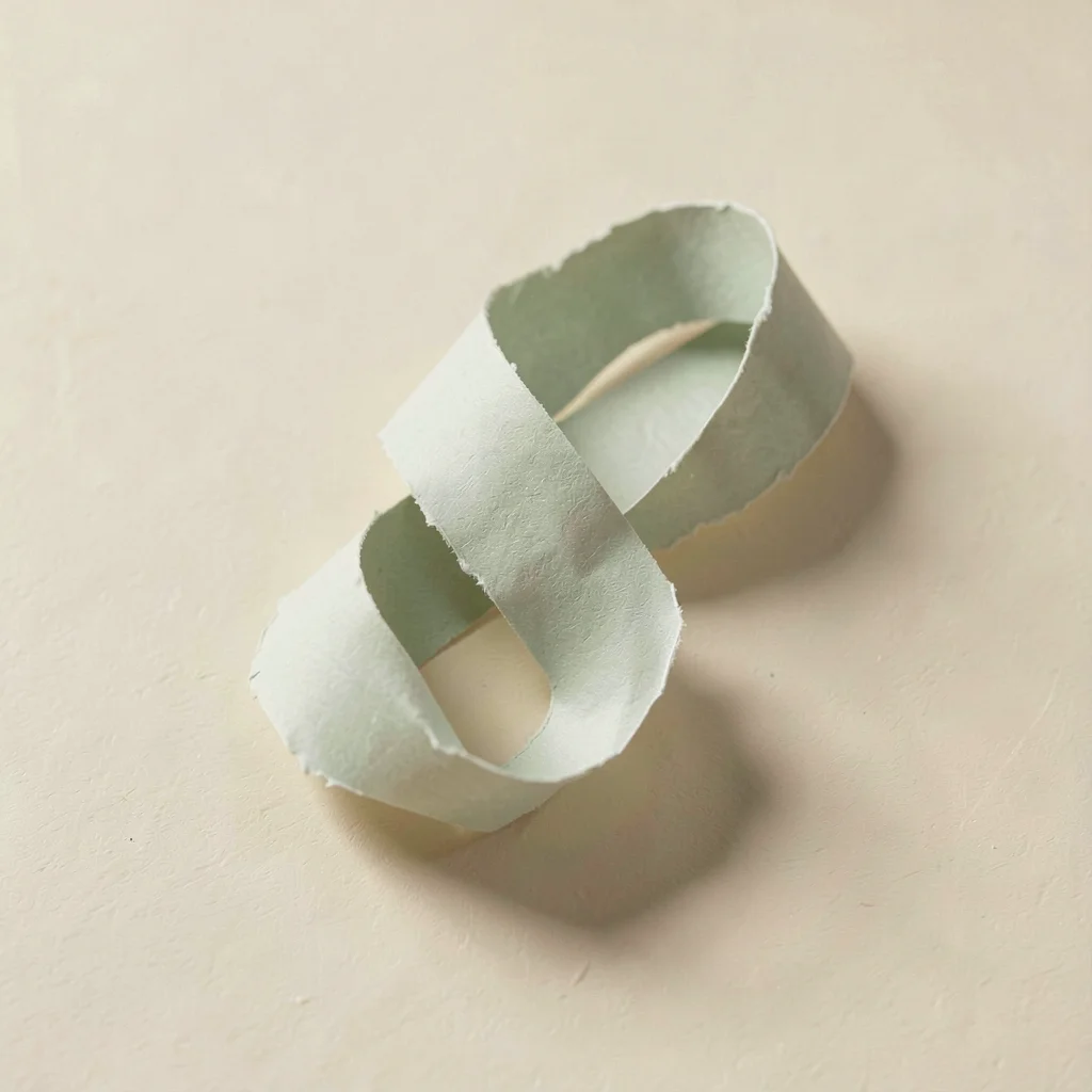Knotted paper sculpture made from one continuous sage ribbon rests on a warm cre