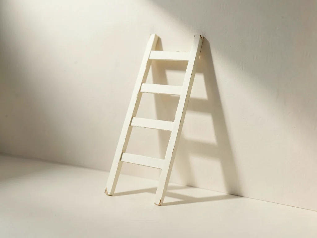 Cream paper ladder leans against an empty papercraft wall casting a long soft ti