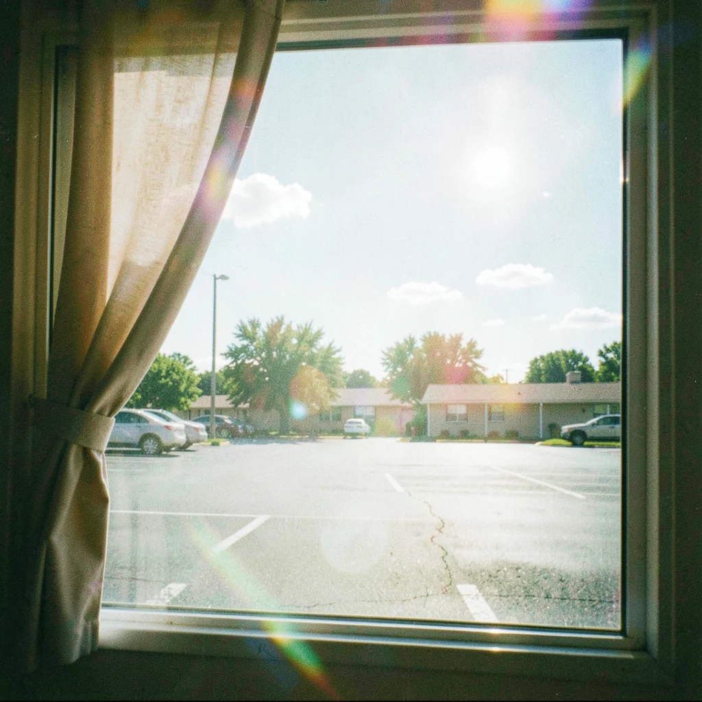 Curtain of a motel window pulled half open onto a midday parking lot with two sl