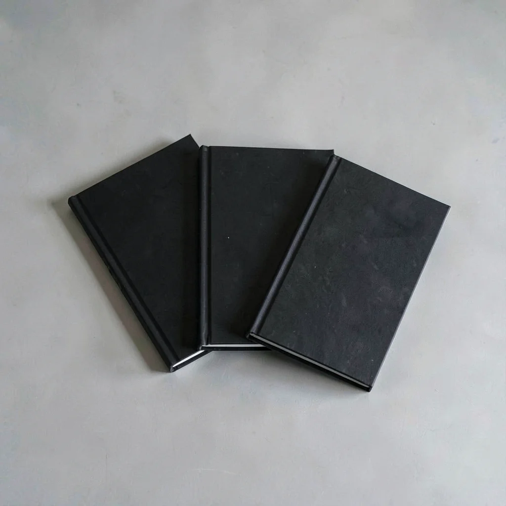 Minimal still of three identical black notebooks fanned on a pale ash desk, hair