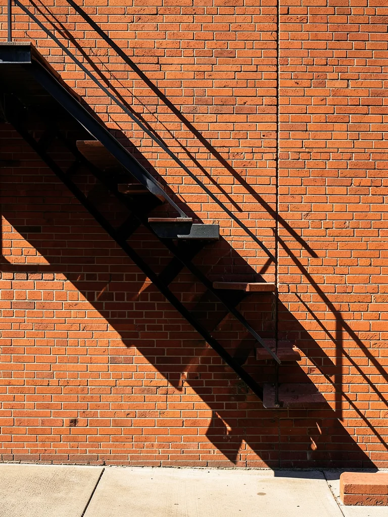 The shadow of a fire escape zigzagging down a brick wall at 10am, creating a per
