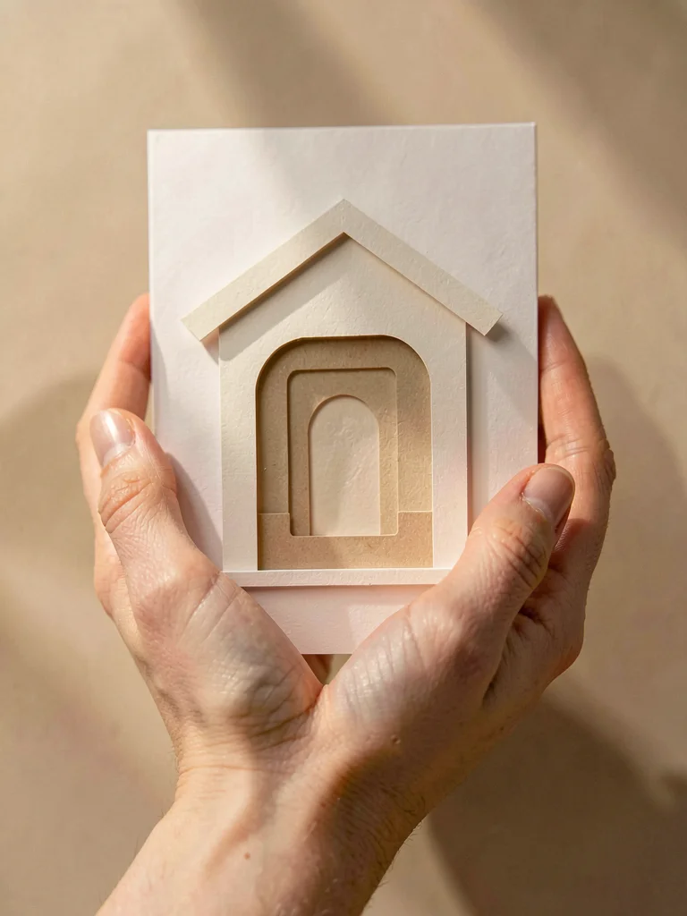 Papercraft brand-card of layered paper-cut hands cradling a paper-cut hall, soft