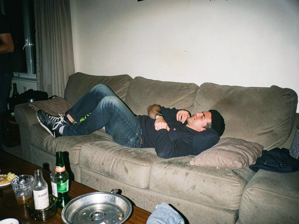 Living room at 7am after a party, someone asleep on couch still in shoes, ashtra