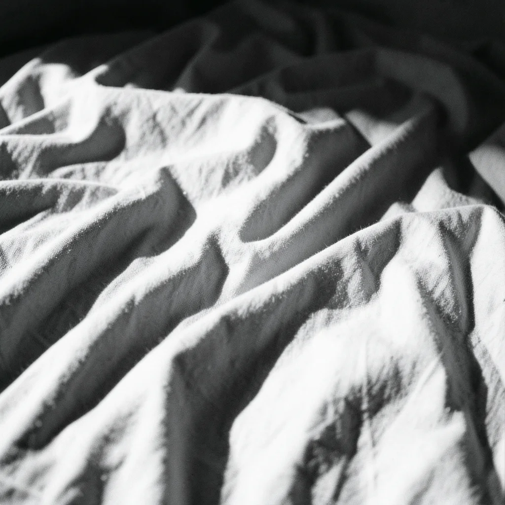 A crumpled bedsheet in morning light, the folds creating a landscape of shadow a