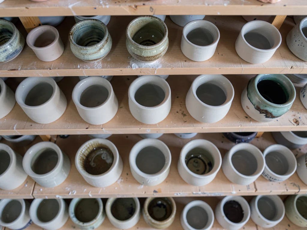 An overhead view of a pottery studio shelf — rows of bisque-fired pieces waiting