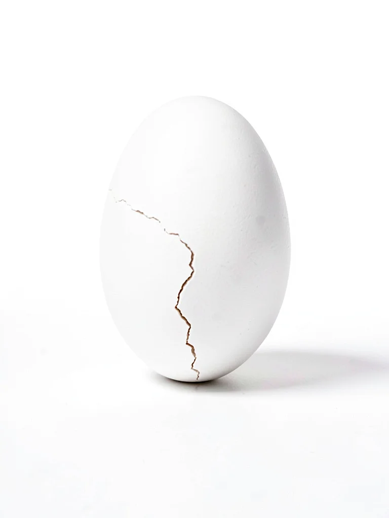 Single white egg balanced on end on white surface, hairline crack just visible, 