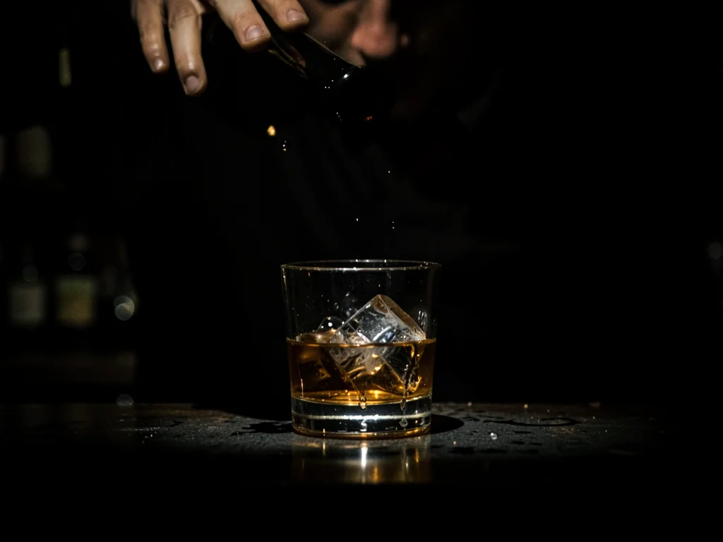 Glass of whiskey on bar, ice melting, single hard light from above, bartender's 