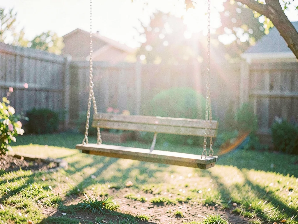 Empty wooden swing still moving in a backyard, late afternoon shadows stretching
