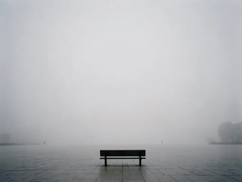 Single bench at frame edge surrounded by vast empty plaza in fog