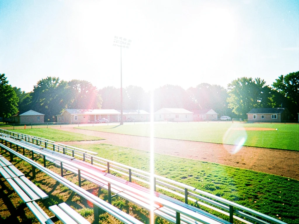 Empty bleachers of a small-town baseball field bathed in light too white for any
