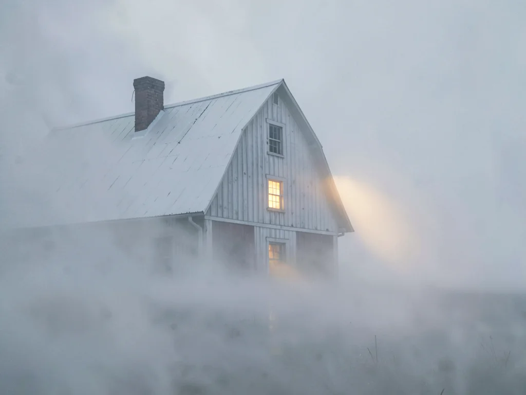 Drifted barn shoulder-deep in white, only roof and chimney visible, a far warm w