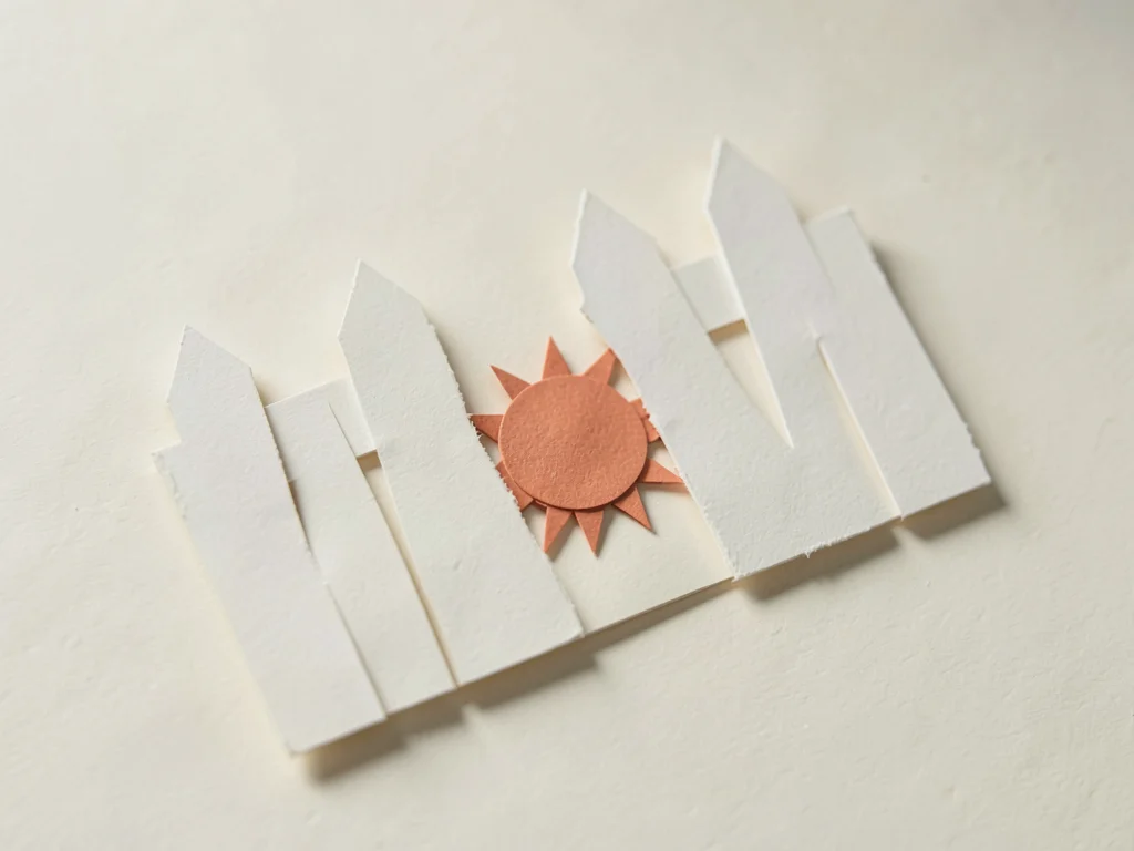Flat papercraft fence with one missing slat reveals a small terracotta paper sun