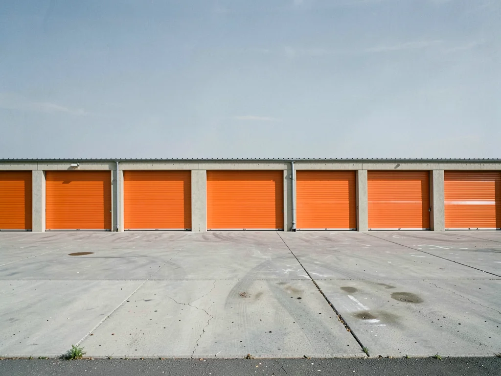 Storage unit facility — repeating orange roll-up doors, concrete drive, flat sky