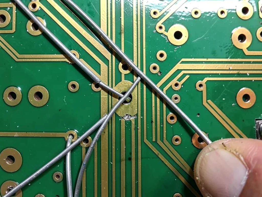 A repair technician's PCB with jumper wires bridging cut traces — silver wires o
