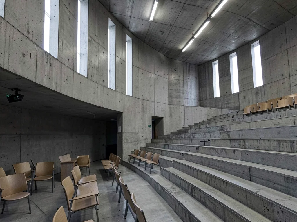 A brutalist university lecture hall — raked concrete tiers, empty chairs, high s