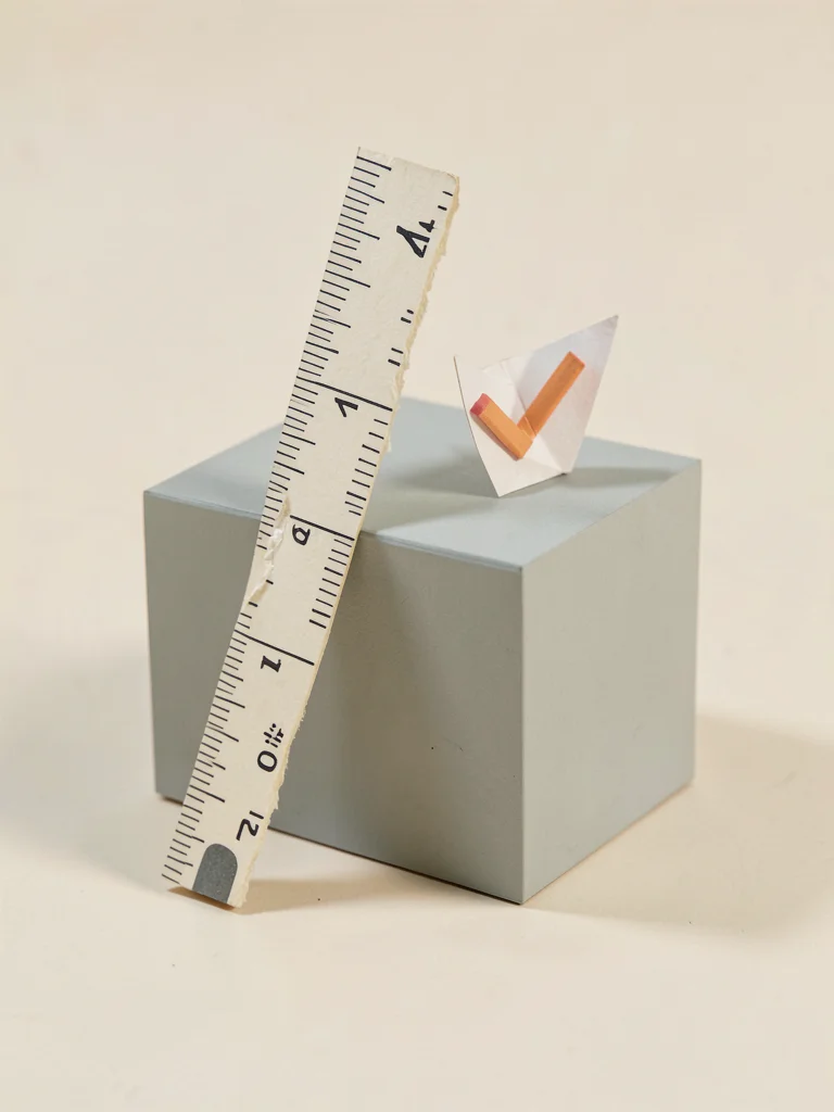 A hand-cut paper measuring stick leaned against a low matte cube, a tiny pencil-