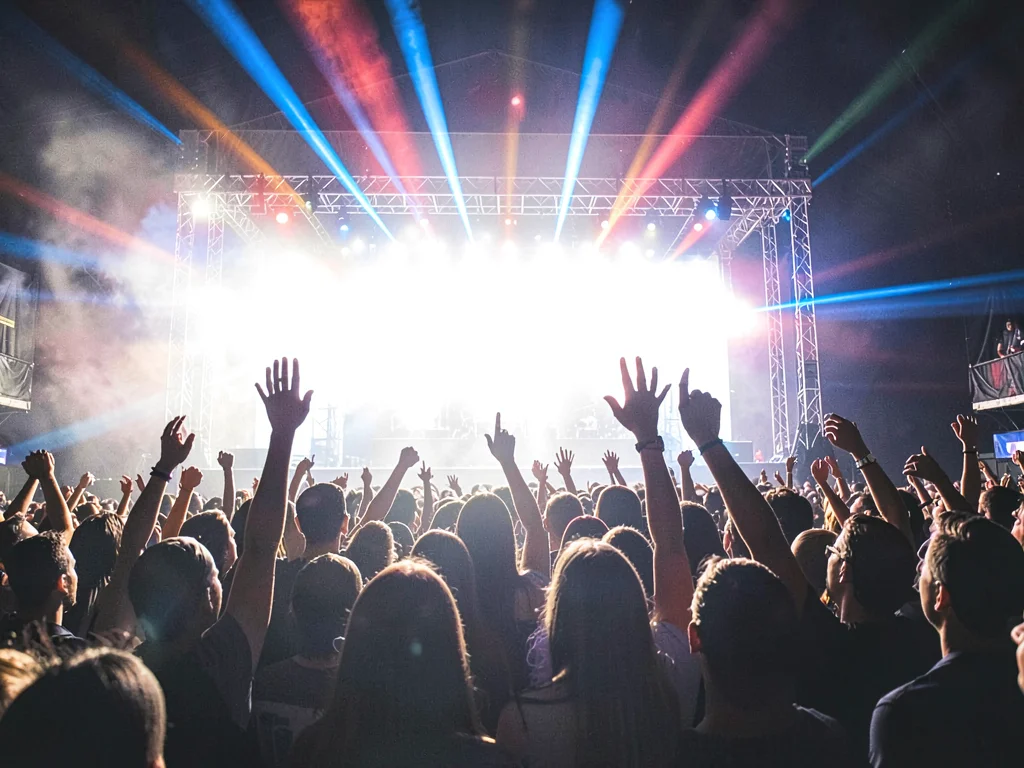 Concert crowd backlit by massive stage lights, raised hands silhouetted