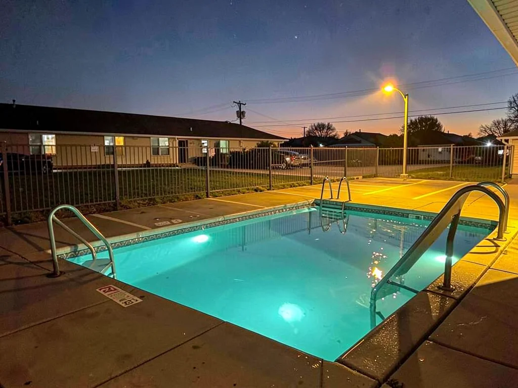 Empty motel pool at dusk, underwater lights creating cyan glow, parking lot ligh