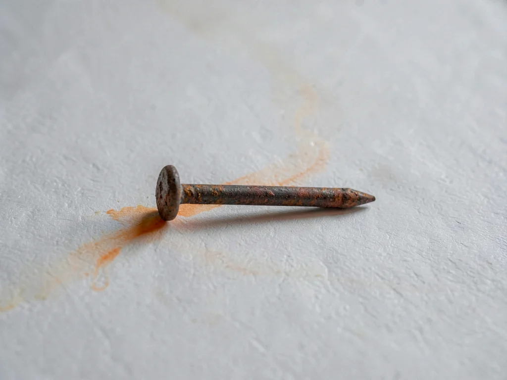 A solitary rusted nail laid horizontal on white-grey paper, oxide bloom blooming