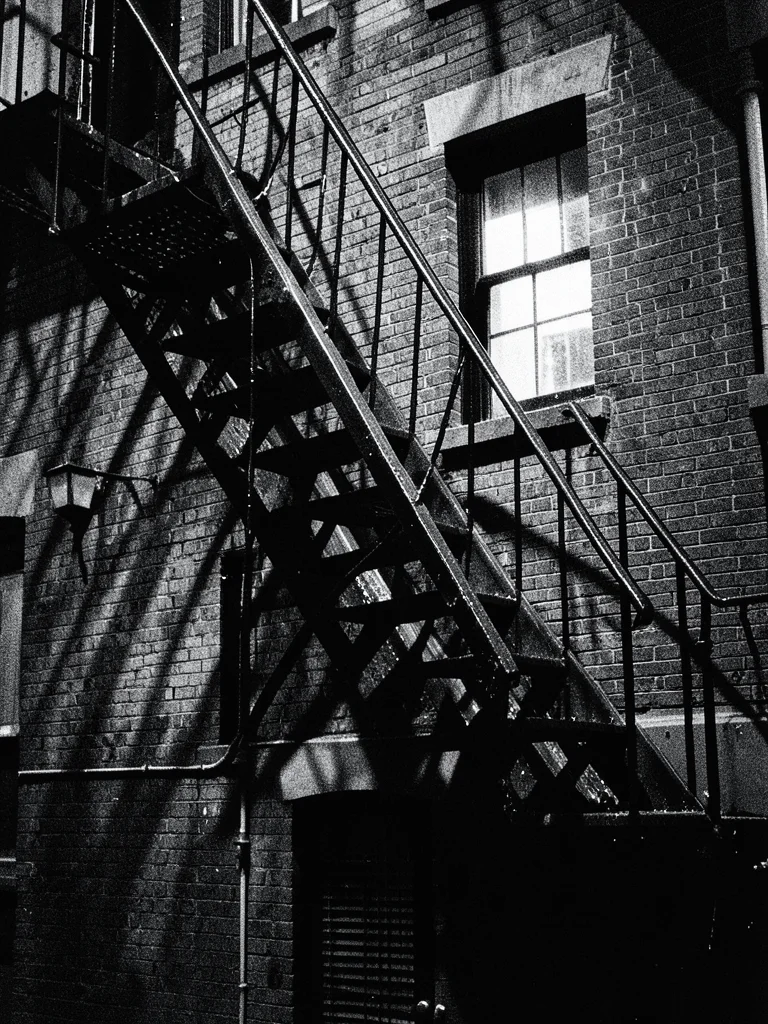 Fire escape zigzagging down a brick building, single lit window, noir shadows