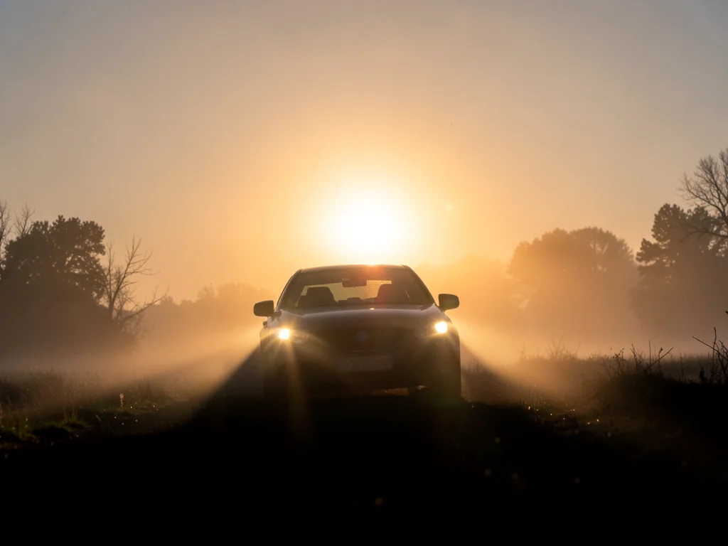 Headlights and fog at dawn shot into the rising sun, a car's silhouette barely v