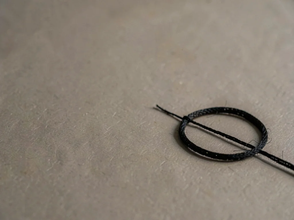 A single coil of black silk cord resting low-right on warm-grey paper, fibres ca