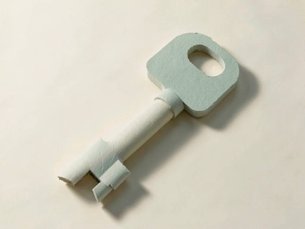 A giant matte-paper key the size of a small house rests on a flat cream surface 