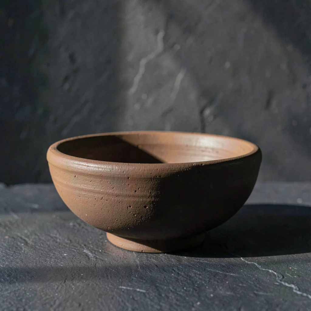 Slate background with a single unfired clay bowl, raking light, deep mineral pal
