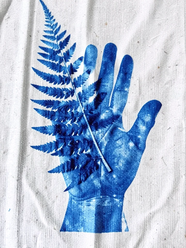 A botanical fern frond overlapping a hand print — nature and body in cyanotype b