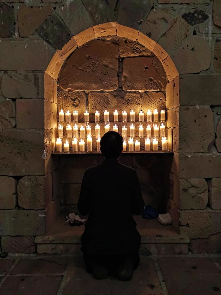 Rows of prayer candles in stone alcove, single figure kneeling in silhouette