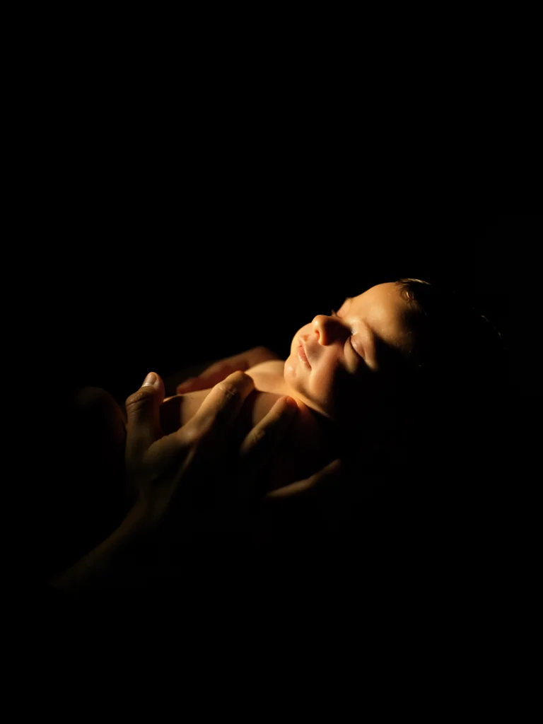 Hands cradling a newborn, a single soft light from a window illuminating the bab