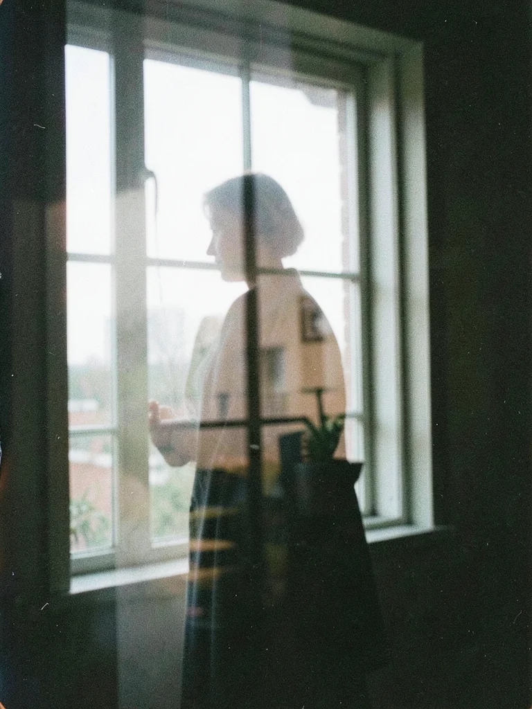 A figure standing at a window, double-exposed with the view beyond, inside and o