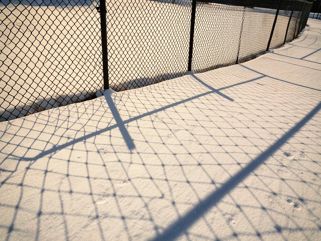 A chain-link fence shadow cast onto fresh snow in low winter afternoon light, th