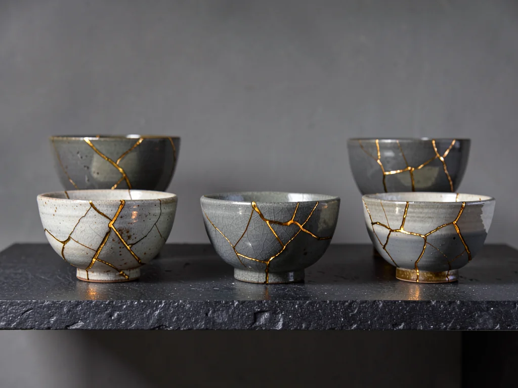 Collection of five repaired bowls on dark stone shelf — varying crack patterns