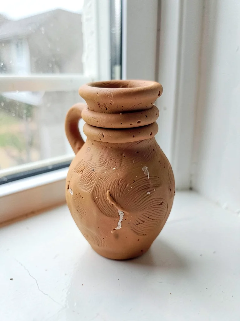 Unfired ochre vase shape with a slumped neck and thumb-pressed coil rings, on no