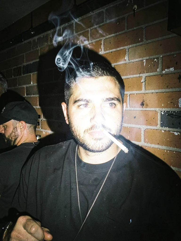 Bouncer's face half-shadowed outside a club at 3am, cigarette smoke frozen mid-e